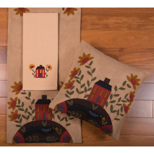 Two square pillow covers with a country house and flowers design, in shades of beige, brown, and red, placed on a wooden floor.