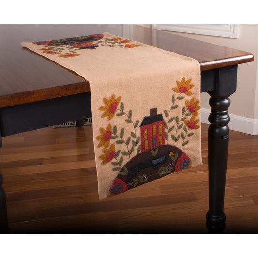 A beige table runner with a floral pattern and a small house design, laid out on a wooden table.