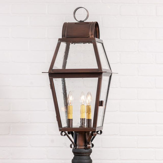 An antique copper outdoor post light with a solid copper structure and glass panels, featuring three light bulbs inside, mounted on a white brick wall.
