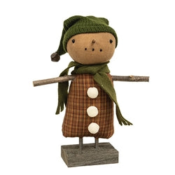 A plush doll resembling a snowman with a brown plaid dress, white pom buttons, a green scarf, and a winter hat, set on a wooden base.
