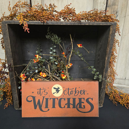Decorative sign with 'It's October Witches' text on a wooden surface with foliage.