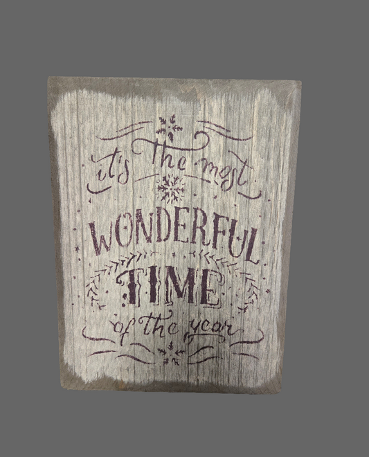 A wooden sign with the phrase 'It's The Most Wonderful Time Of The Year' written in a decorative font, accompanied by floral designs and stars.