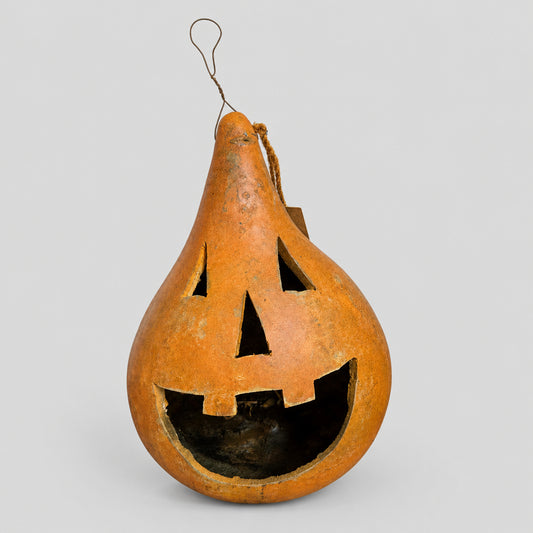 An orange carved gourd lantern with a hanging loop, photographed against a rustic whitewashed wooden wall and dark tabletop.