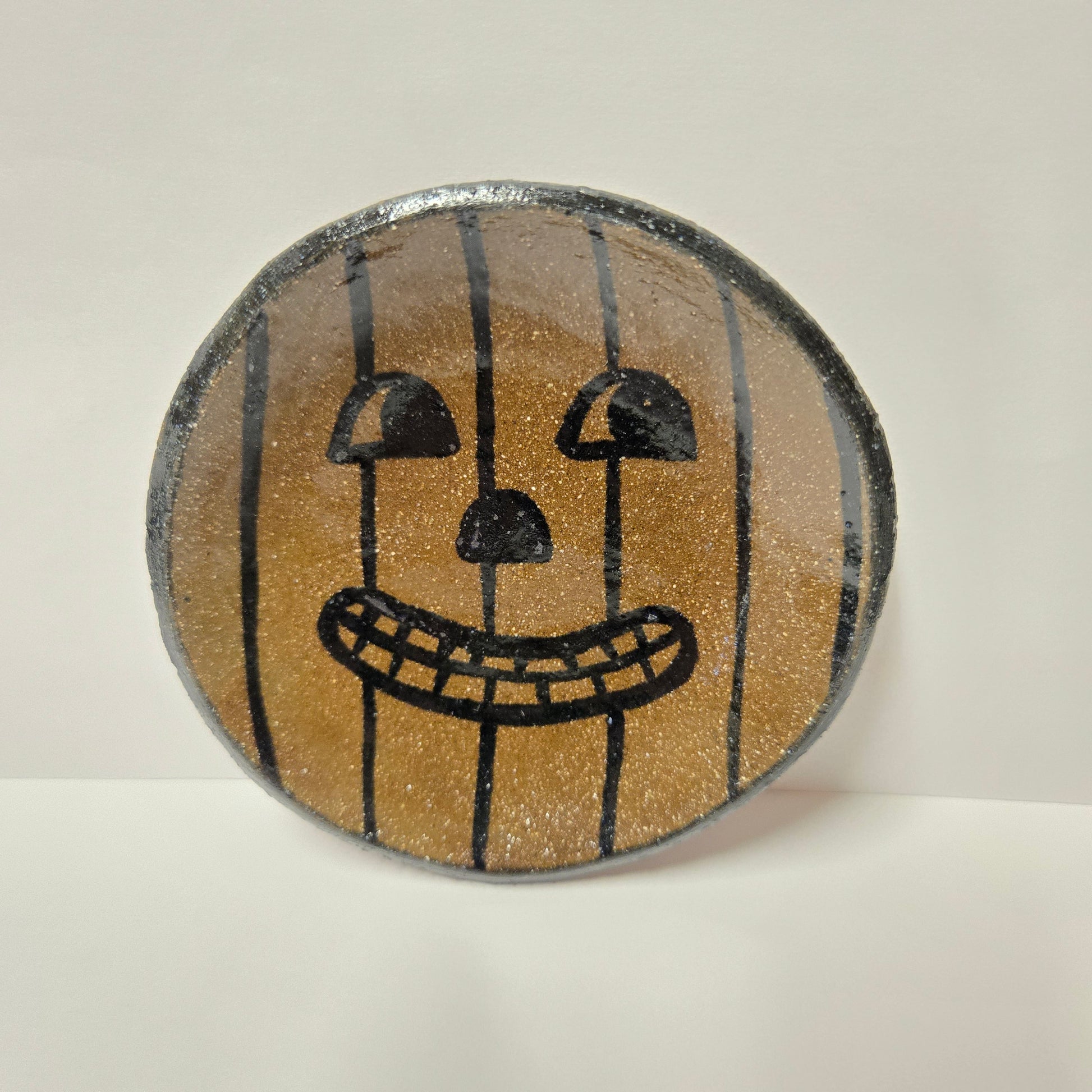 A round, hand-painted stoneware plate featuring a jack-o'-lantern face design on a glittery gold background with black vertical stripes and black facial features, set against a plain off-white background.