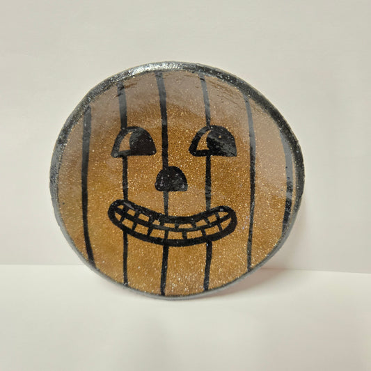 A round, hand-painted stoneware plate featuring a jack-o'-lantern face design on a glittery gold background with black vertical stripes and black facial features, set against a plain off-white background.