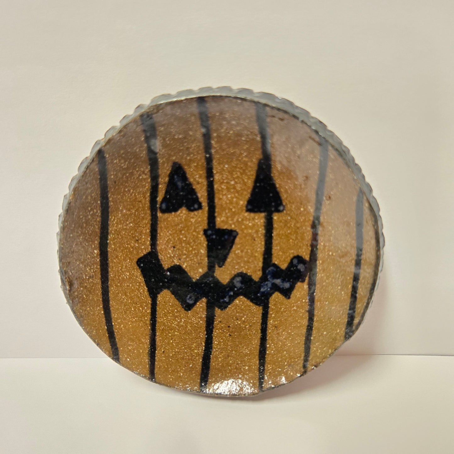 A round, handcrafted decorative plate with a glittery gold-orange pumpkin design, featuring a black hand-painted jack-o'-lantern face and vertical black stripes, set against a plain off-white background.