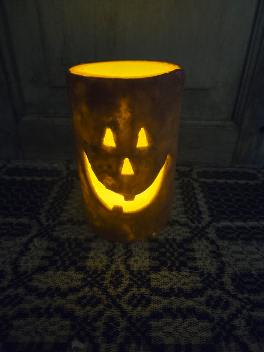 A battery-operated pillar candle with a jack-o'-lantern design, featuring a grungy wax coating and a toothy jack-o'-lantern face pressed into the wax, illuminated from within.