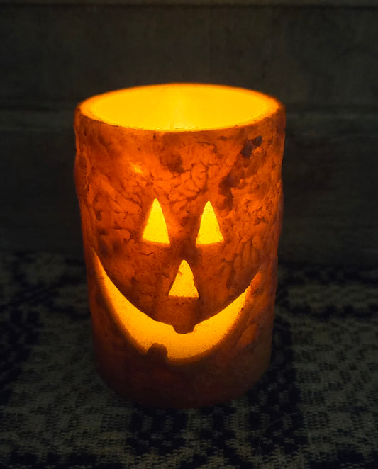 A battery-operated orange pillar candle with a carved jack-o'-lantern face that is glowing.