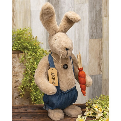 A plush toy depicting a bunny doll wearing denim overalls and holding an orange carrot. The doll has a light tan color and features a jingle bell necklace with flexible ears and flat feet for standing.