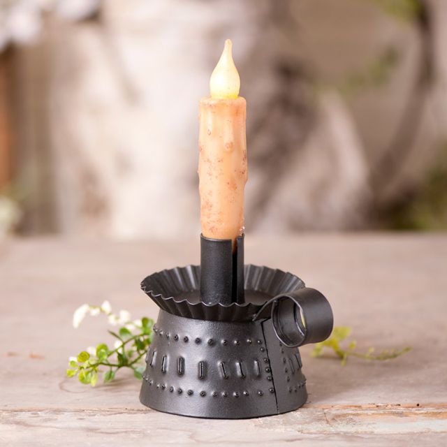 A black metal candle holder with a smokey finish, designed to hold a candle, placed on a wooden surface with a floral decoration beside it.