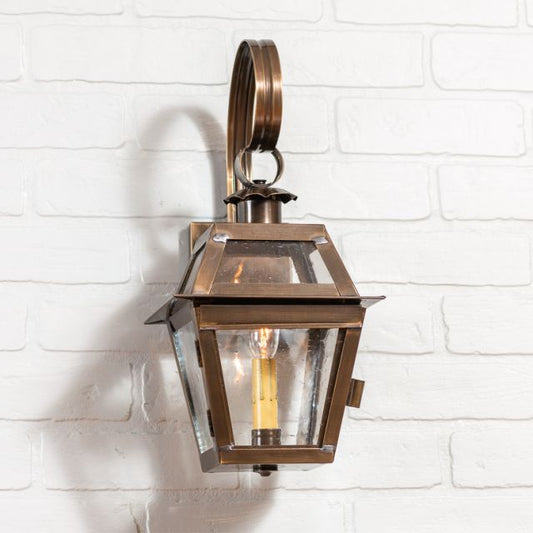 A solid weathered brass outdoor wall light fixture with a clear glass cover and a visible bulb inside, mounted on a white brick wall.