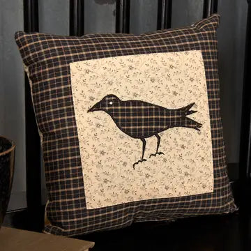 A square decorative throw pillow with a rustic plaid border in dark brown and beige, featuring a central cream-colored patch with a black silhouette of a raven.