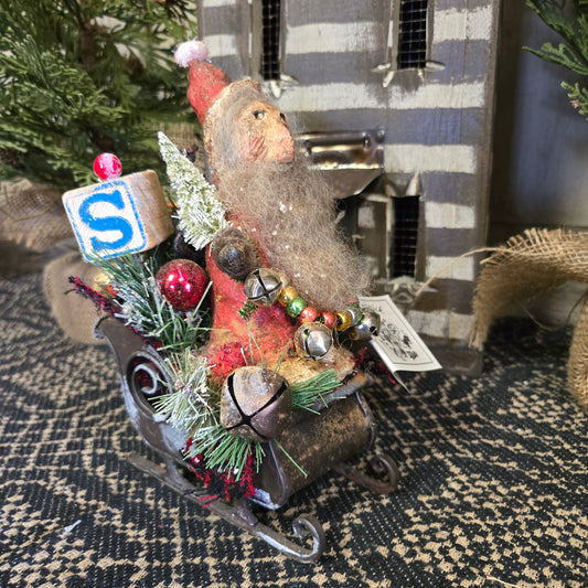 A decorative figurine of Santa Claus sitting in a metal sleigh, adorned with colorful Christmas decorations, placed on a patterned surface.