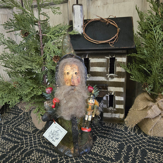 Handcrafted Santa figurine made of chalkware, depicted holding a bell and a nutcracker, displayed among greenery.