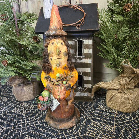 Handcrafted figurine of a witch with a basket, detailed with a striped hat and a smiling face, placed on a textured surface with holiday decorations in the background.