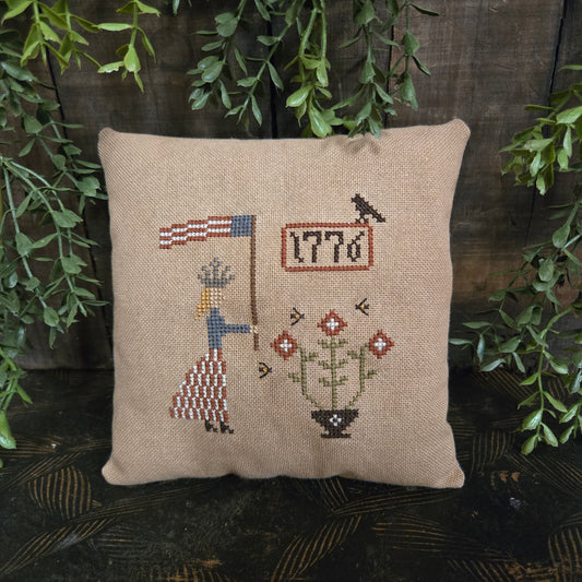 Decorative pillow with cross-stitch design on a wooden surface with greenery