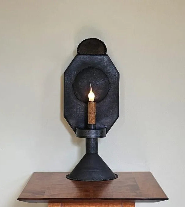 A black tin table light with a candle-like light at its center, placed on a wooden surface, against a plain wall background.