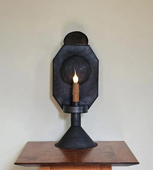 A black tin table light with a candle-like light at its center, placed on a wooden surface, against a plain wall background.