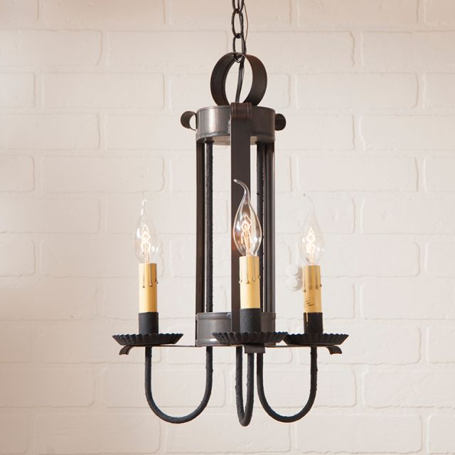 Black metal chandelier with three candle-style light fixtures against a white brick wall.