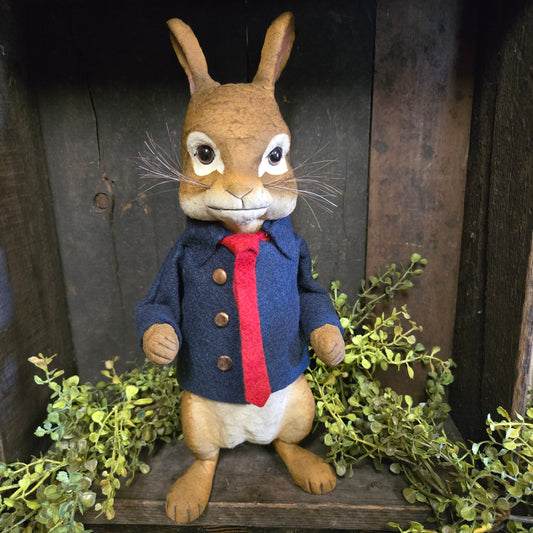 Stuffed animal resembling a rabbit in a blue outfit with a red collar on a wooden surface with greenery.
