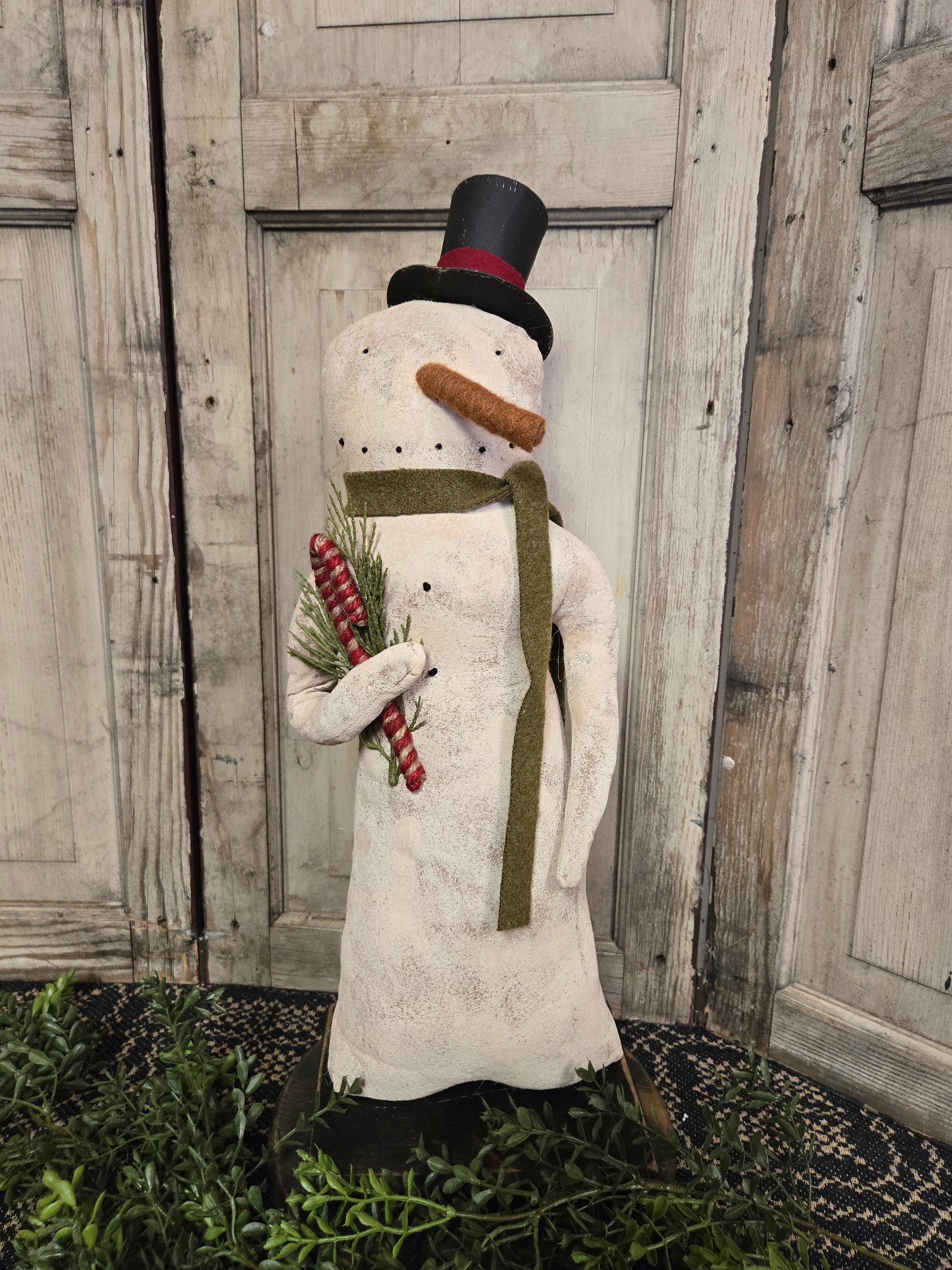 White snowman with top hat and green scarf holding a candy cane standing in front of a wood door