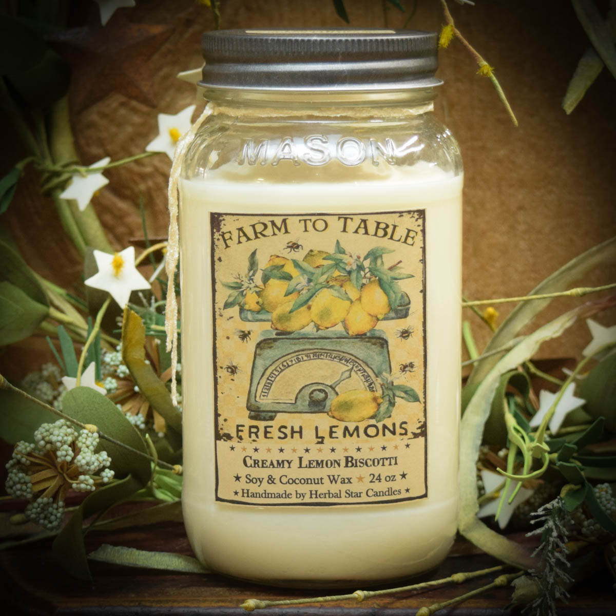 A mason jar candle labeled 'Farm to Table' with 'Fresh Lemons' and 'Cream Lemon Biscotti' scents, made with soy and coconut wax, displayed on a table with decorative flowers and a herbal star card in the background.