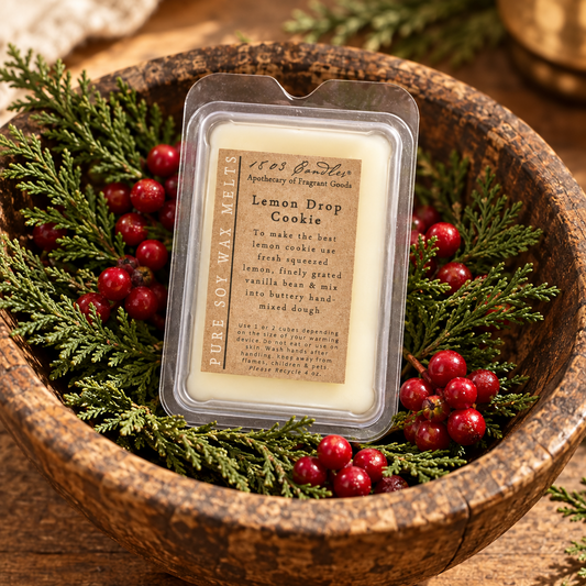 A packaged soy wax melt labeled 'Lemon Drop Cookie' by 1853 Garden Apothecary of Fragrance Co., displayed in a rustic wooden bowl with evergreen branches and red berries.