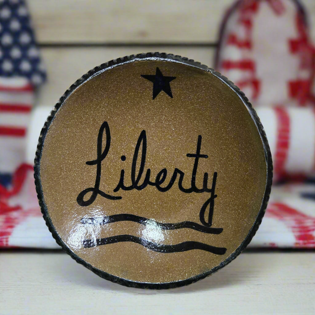 A round stoneware plate with the word 'Liberty' written on it, featuring a star and wavy line design, brown in color with a black rim.