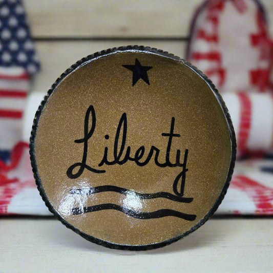 A round stoneware plate with the word 'Liberty' written on it, featuring a star and wavy line design, brown in color with a black rim.