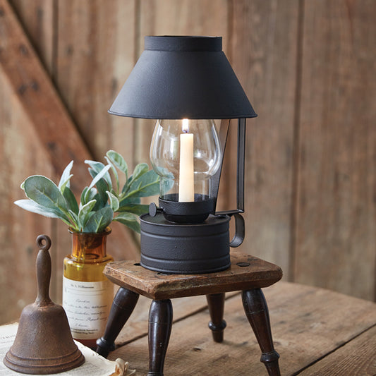 A black lantern-style candle holder with a glass top and a metal base, placed on a wooden surface with a plant and a bell beside it.