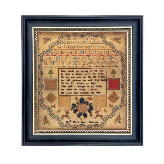 A framed needlepoint sampler print featuring upper and lowercase alphabets, birds, deer, cherubs, dogs, trees, and color blocks, with a visible signature at the bottom.
