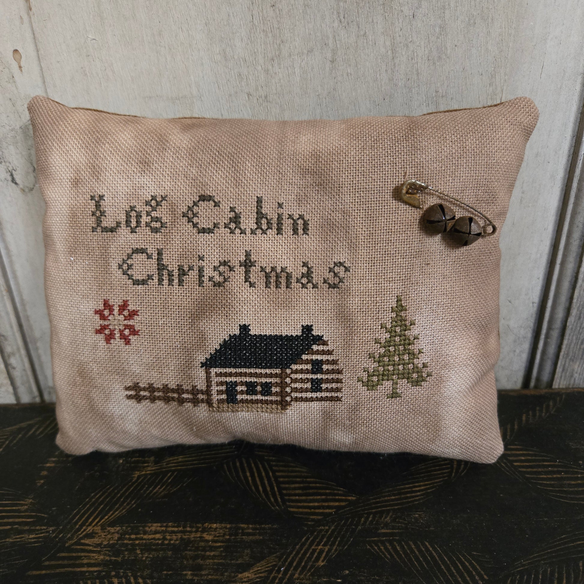 Decorative pillow with cross-stitched design of a cabin and text on a wooden surface.