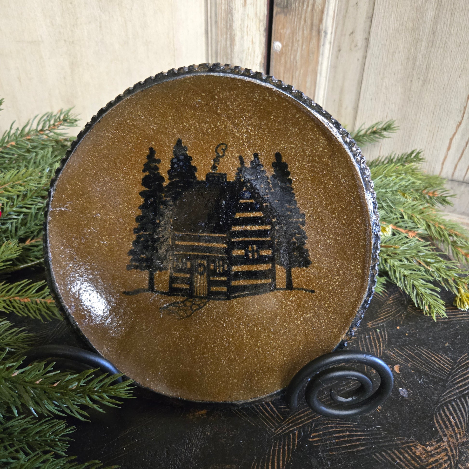 Decorative ornament with a cabin silhouette on a brown background, surrounded by greenery.