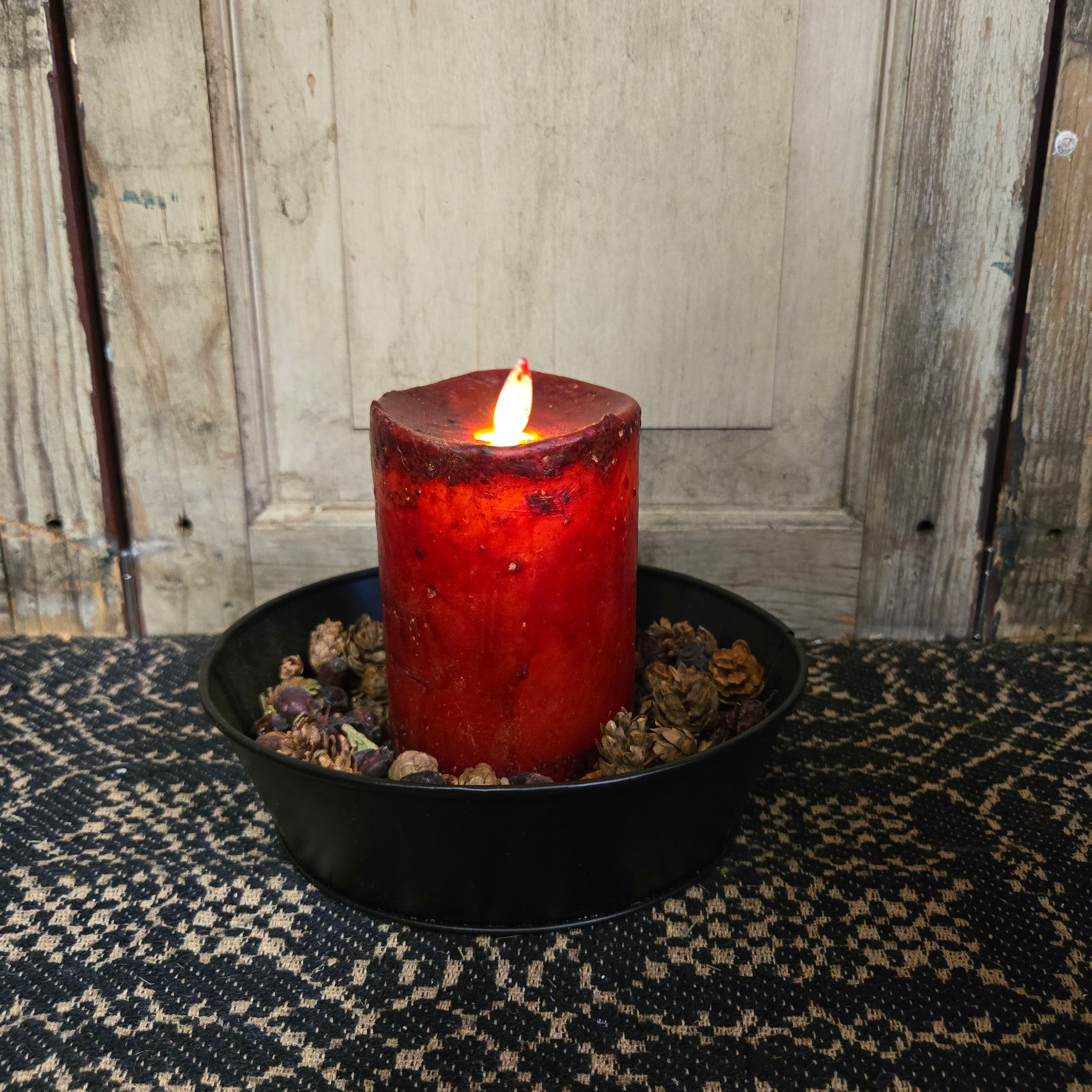 A synthetic moving flame candle in burgundy color appears to be burning, placed on a black dish on a patterned fabric.