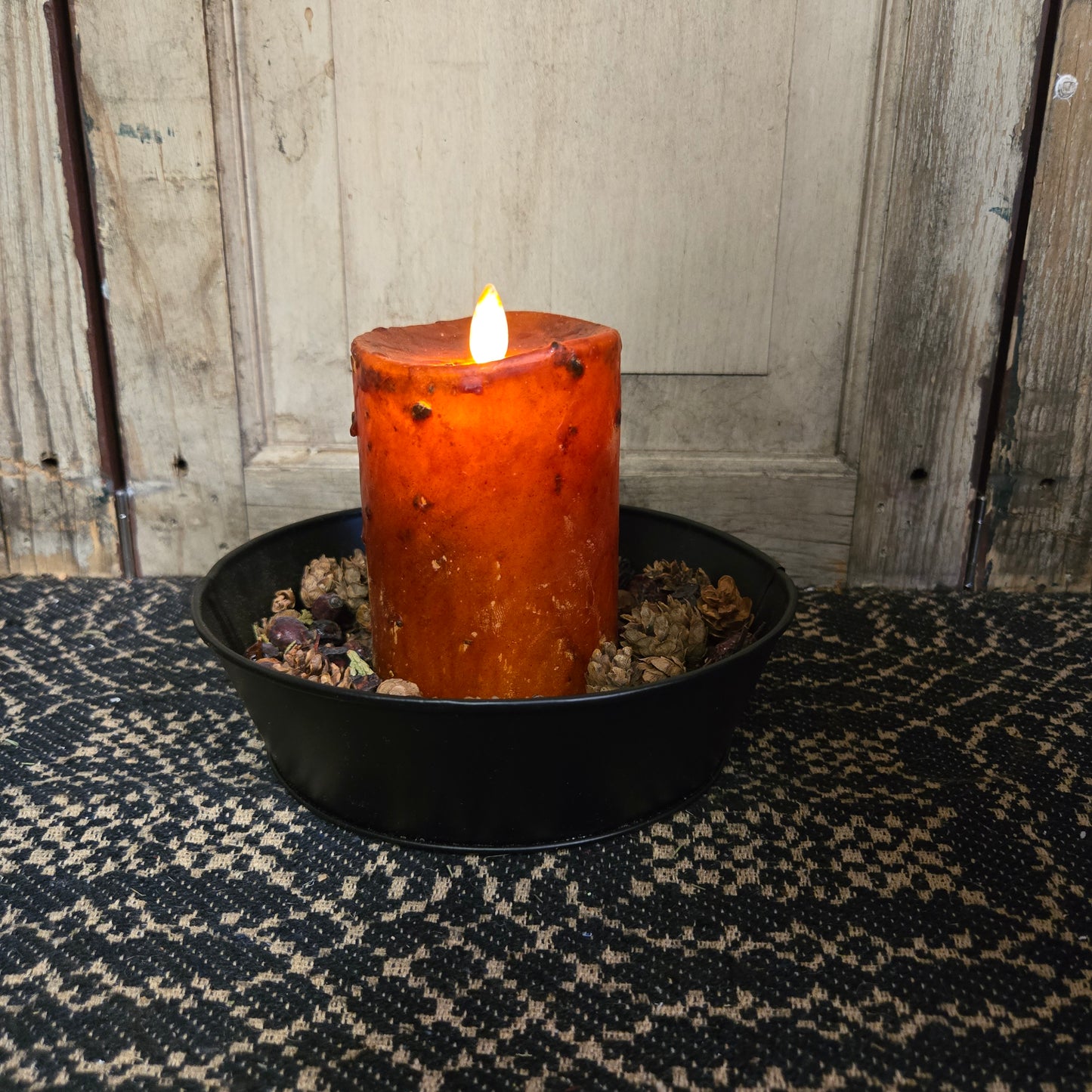 A 3 inch by 4 inch moving flame pillar candle in burnt orange color, placed on a black dish on a lace doily with a wooden background.