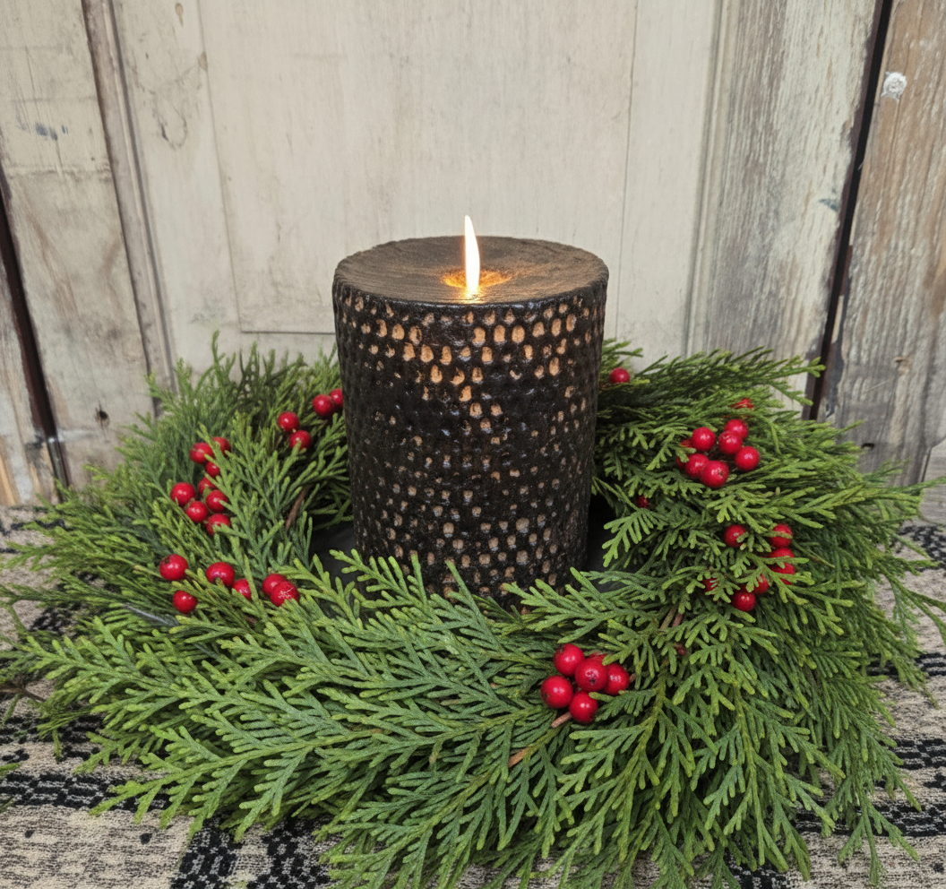 A black beeswax honeycomb pillar candle with a realistic moving flame, placed on a round black base, surrounded by green foliage.