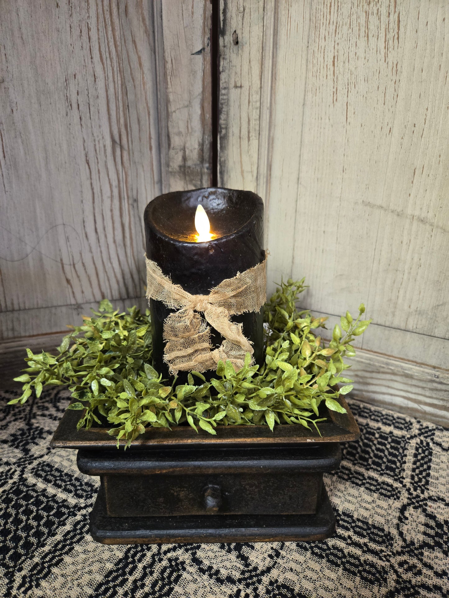 A black 5-inch moving flame pillar candle with a primitive design, placed on a decorative tray surrounded by greenery.