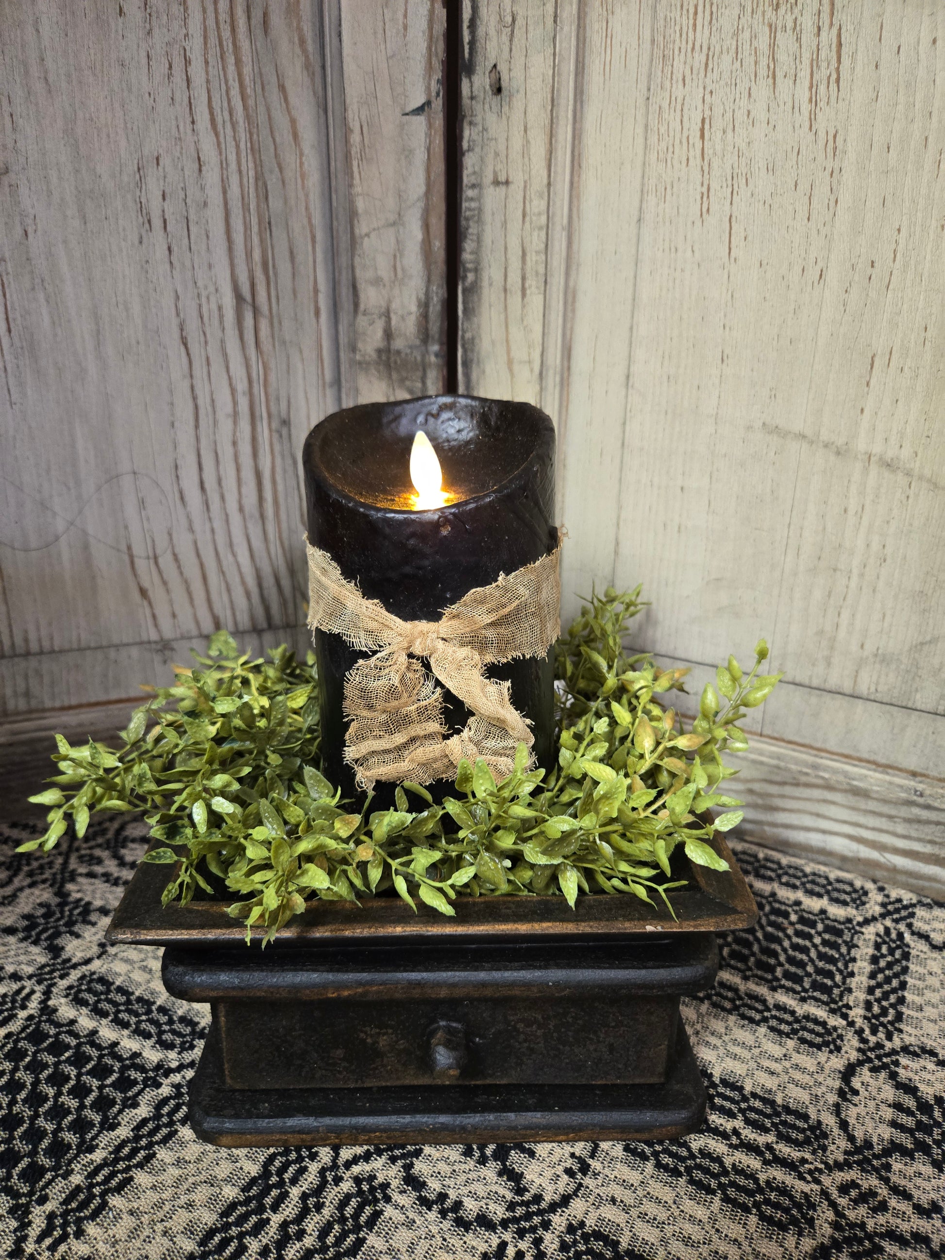 A black 5-inch moving flame pillar candle with a primitive design, placed on a decorative tray surrounded by greenery.