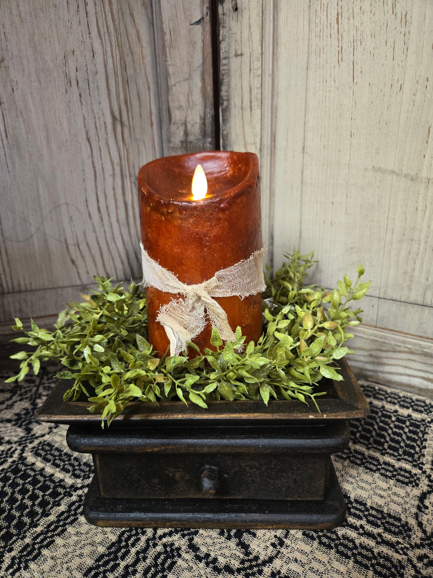 A realistic moving flame 5-inch pillar candle in burnt orange color, presented on a wooden surface surrounded by greenery.