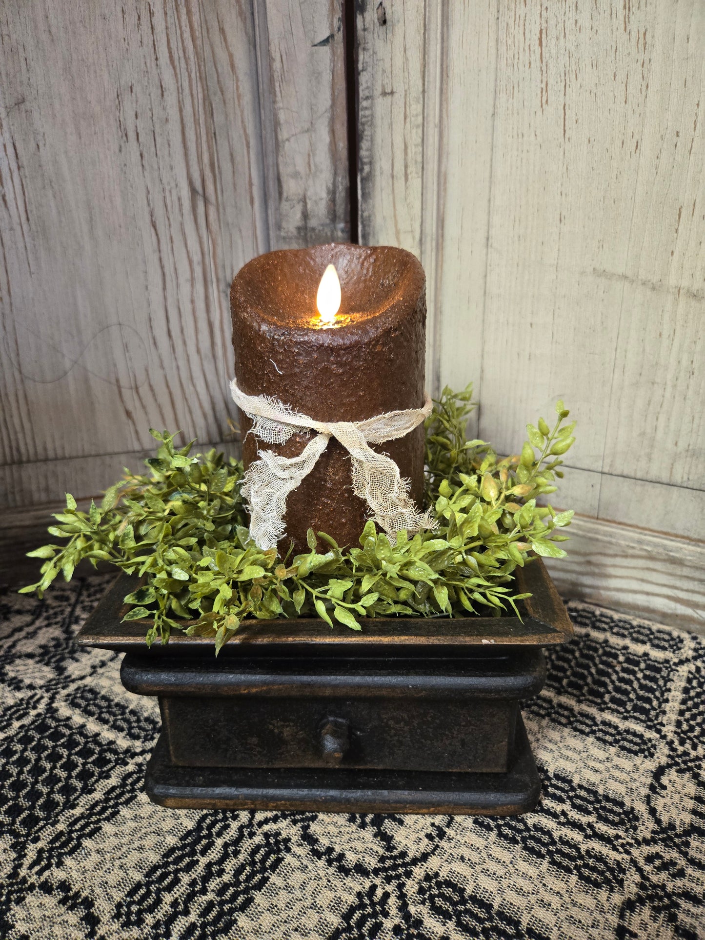 A realistic moving flame 5-inch pillar candle in dark brown with a primitive appearance, placed on a decorative tray surrounded by greenery.