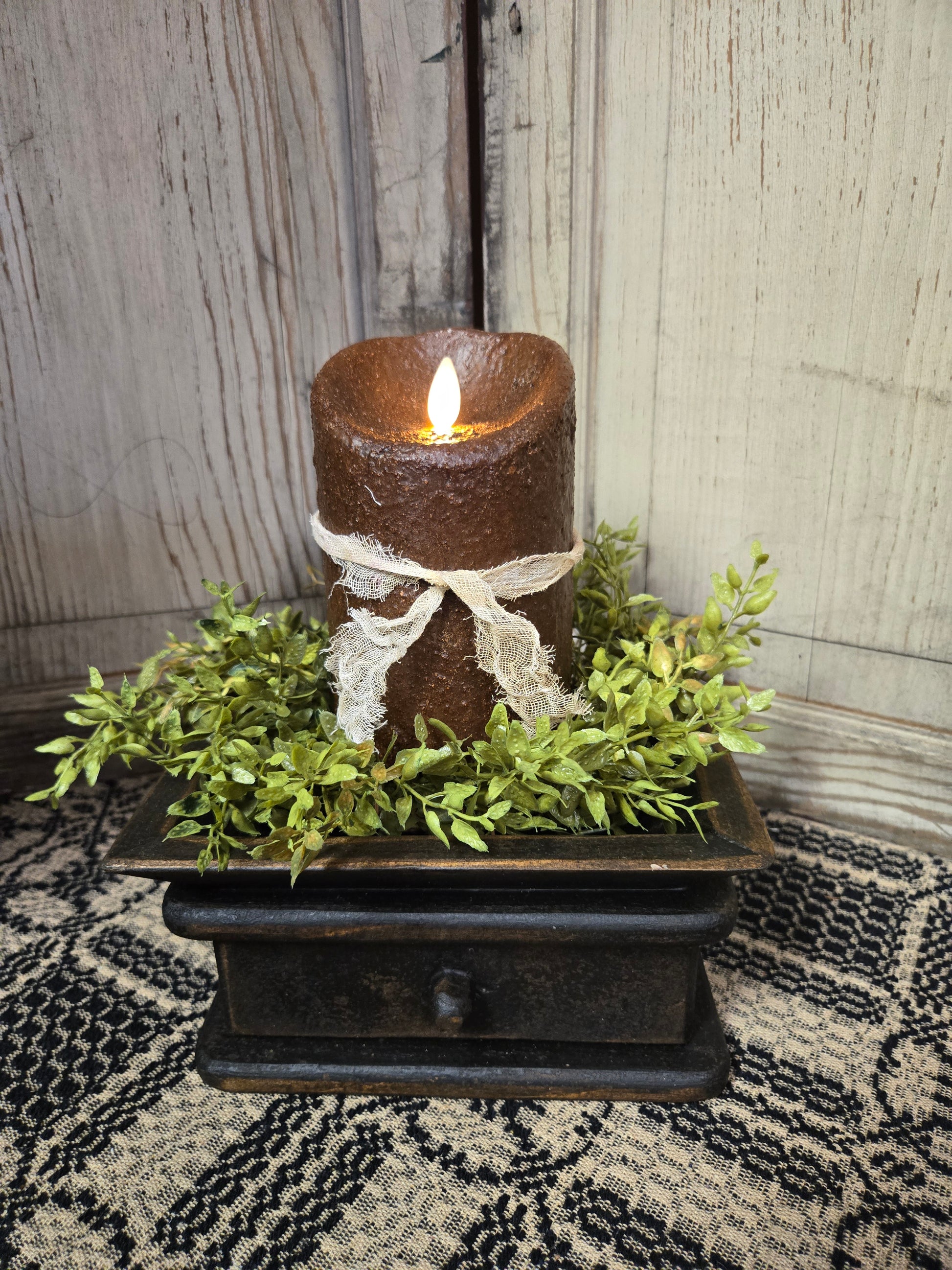 A realistic moving flame 5-inch pillar candle in dark brown with a primitive appearance, placed on a decorative tray surrounded by greenery.