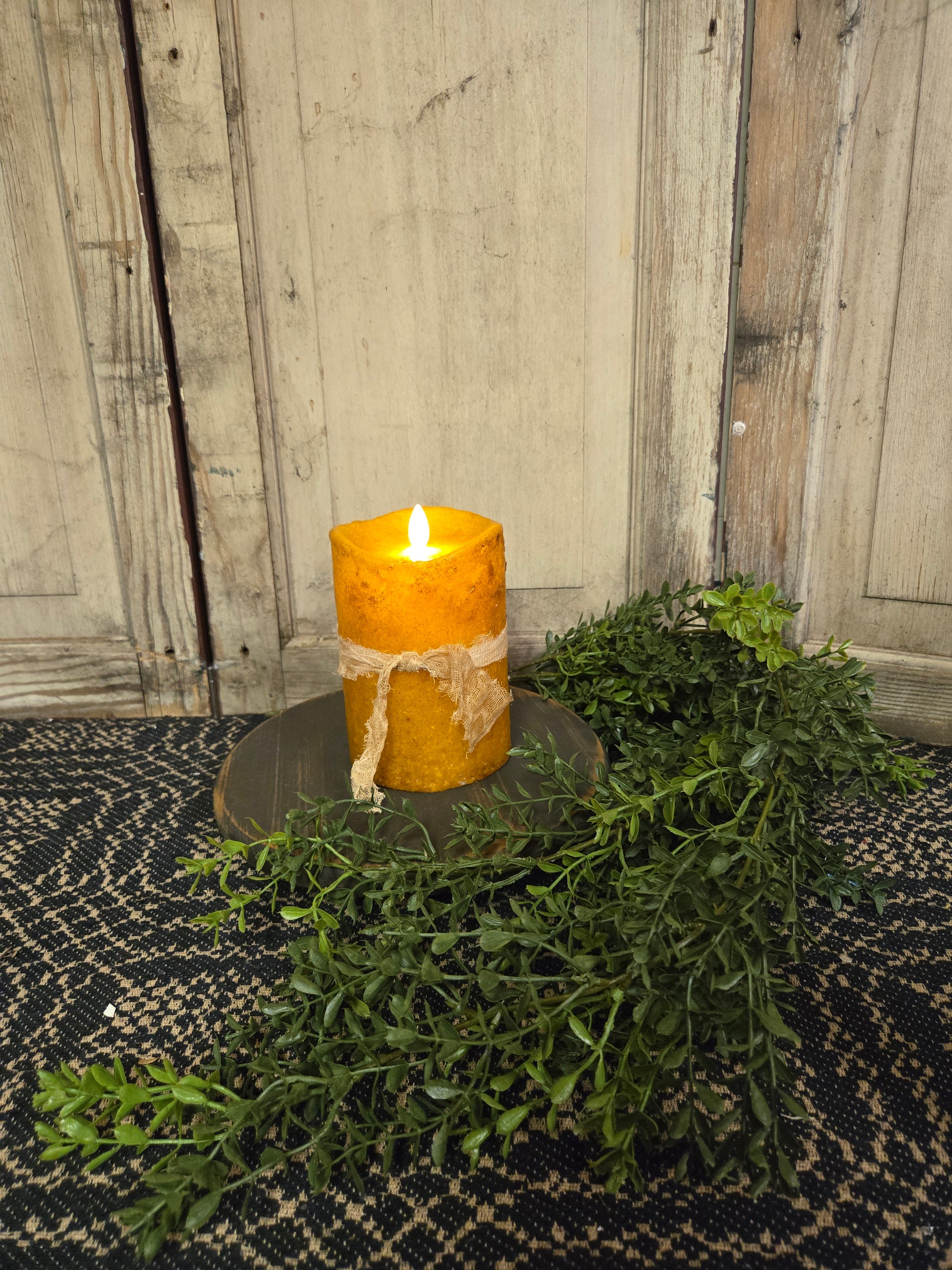 A battery-operated moving flame pillar candle with a yellow flame, surrounded by green foliage and tied with a cheesecloth fabric. Set on a dark textured surface against a wooden backdrop.