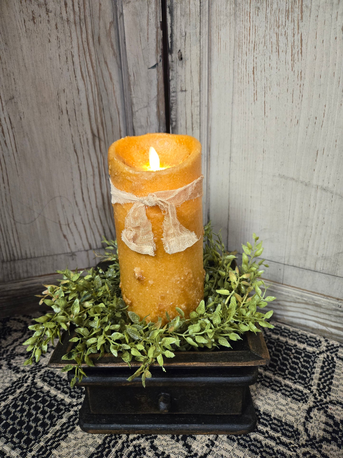 A 7-inch mustard-colored moving flame pillar candle with a primitive, grungy appearance, sitting on a decorative bed of green leaves in a black container. The candle is battery operated with a cheesecloth fabric tie around it.
