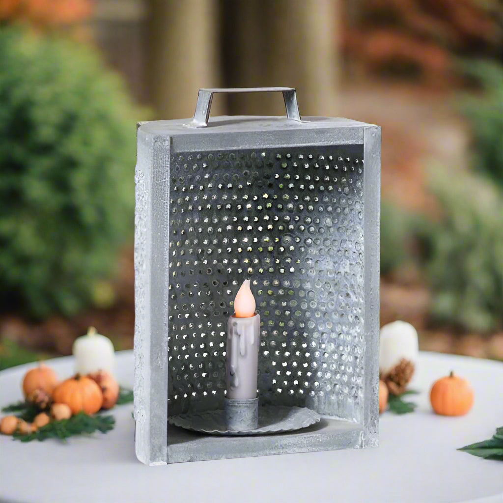 A weathered zinc candle holder with a grid design and a lit candle inside, placed on a white surface with a vintage background.