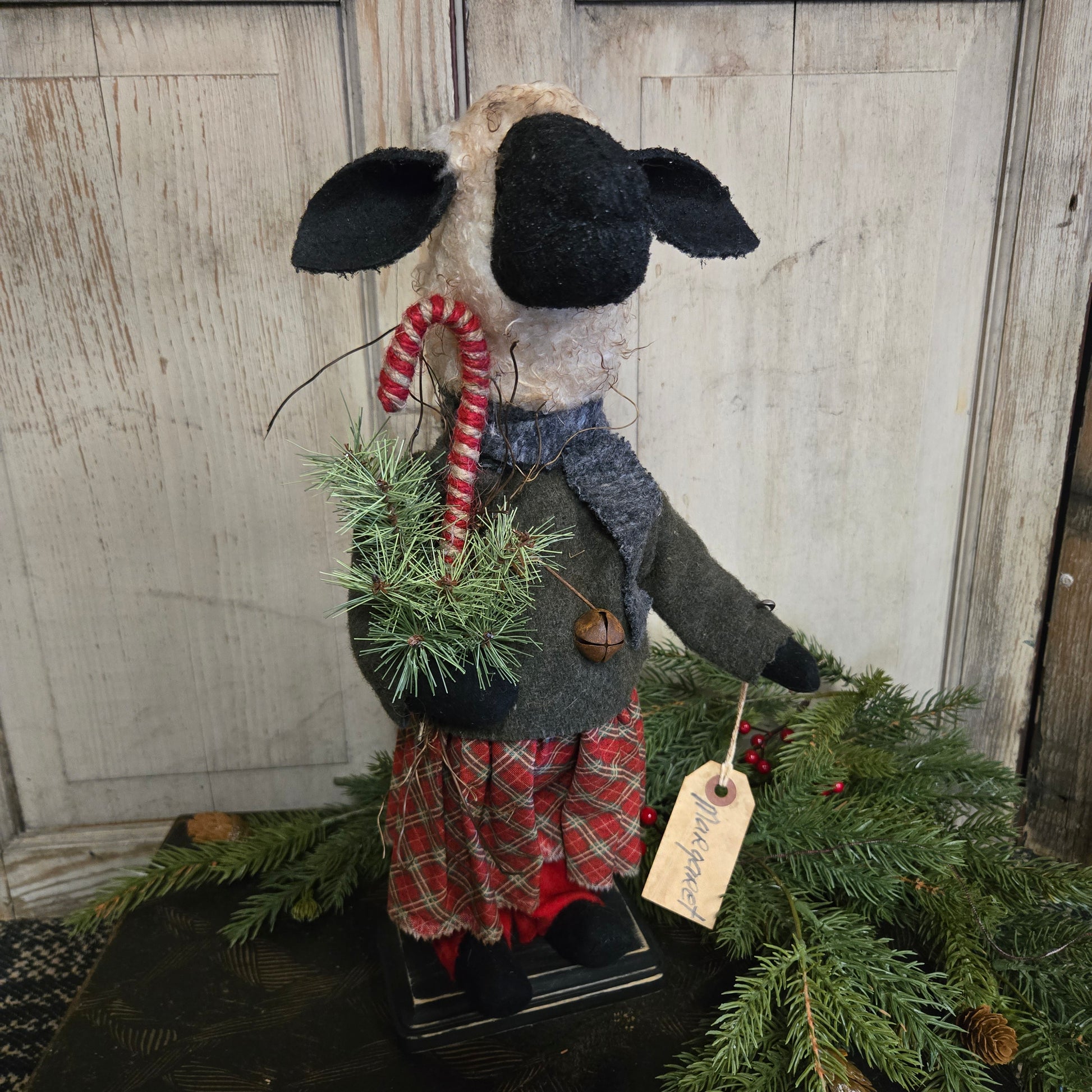 Decorative sheep with a plaid outfit and candy cane, standing next to a Christmas tree on a wooden surface.