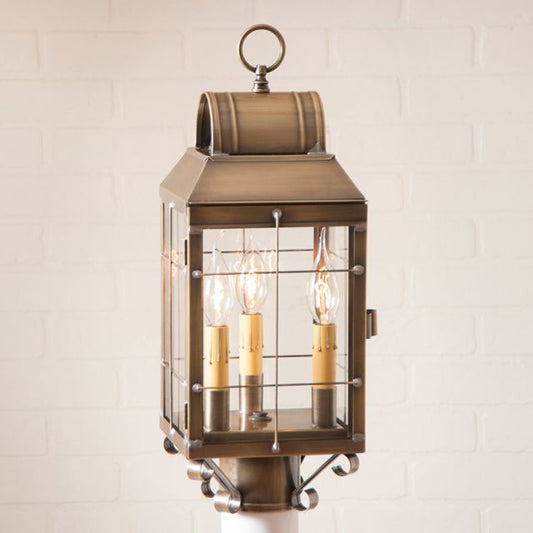 A weathered brass post lantern with clear glass and three internal light bulbs, placed on a white surface with a brick wall in the background.