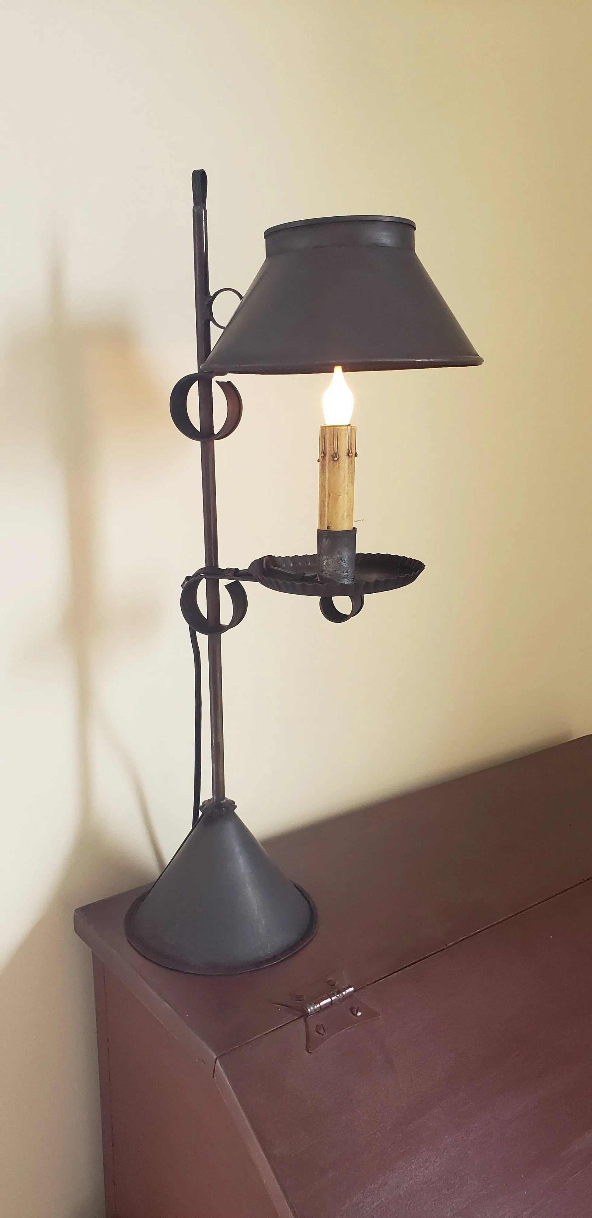 A black Meriden Tin Table Light with a lit candle inside, placed on a table with a shadow falling on the wall.