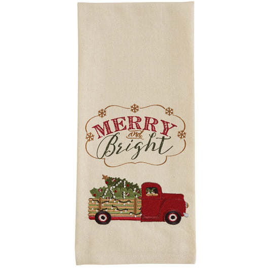 A beige dishtowel with an embroidered red truck carrying Christmas trees and the text 'Merry & Bright'.