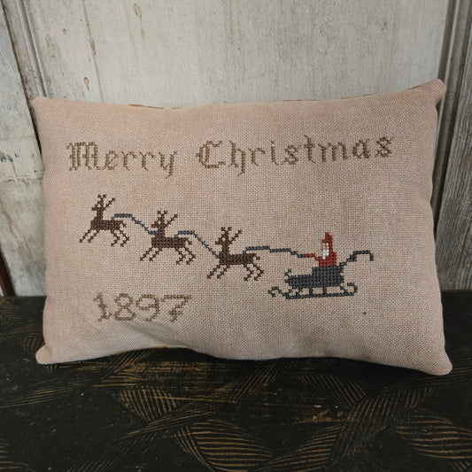Decorative pillow with cross-stitched design of Santa in a sleigh and text 'Merry Christmas 1897' on a wooden surface.