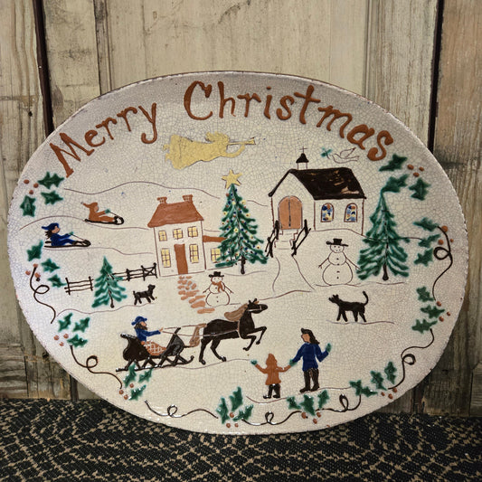 A 16 inch oval ceramic plate with a 'Merry Christmas' message and festive holiday decorations including a cabin, reindeer, and people.